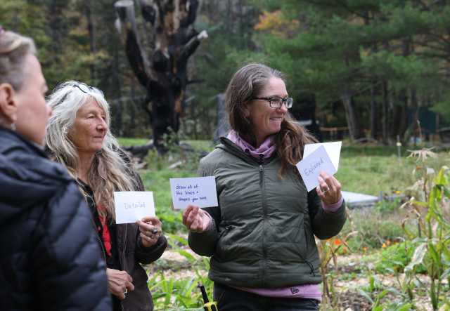Three women stand in a garden space, hold cards that are part of a group activity