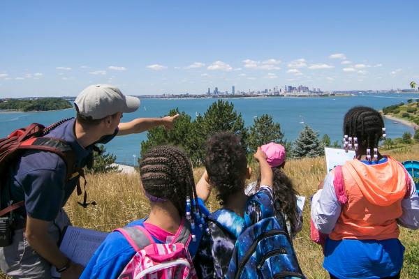 Young students and educator with backs to camera point toward the City of Boston as seen from the Harbor Islands. They stand atop a grassy hill, with ocean surrounding the island.
