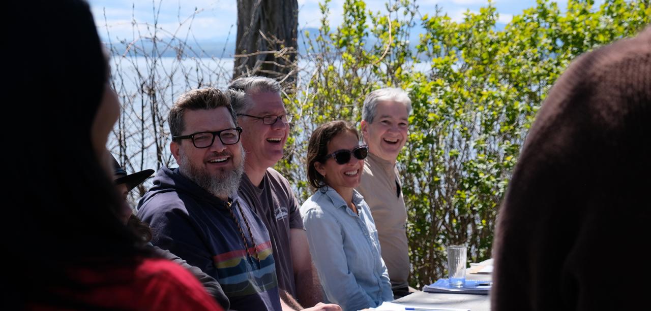 Four educators laugh around a sunny, spring picnic table at lakeshore at Shelburne Farms. Green bushes stand in background with lake and Adirondack Mountains in distance.