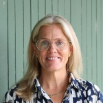 Woman with long blonde hair and glasses smiles at the camera in front of green beadboard