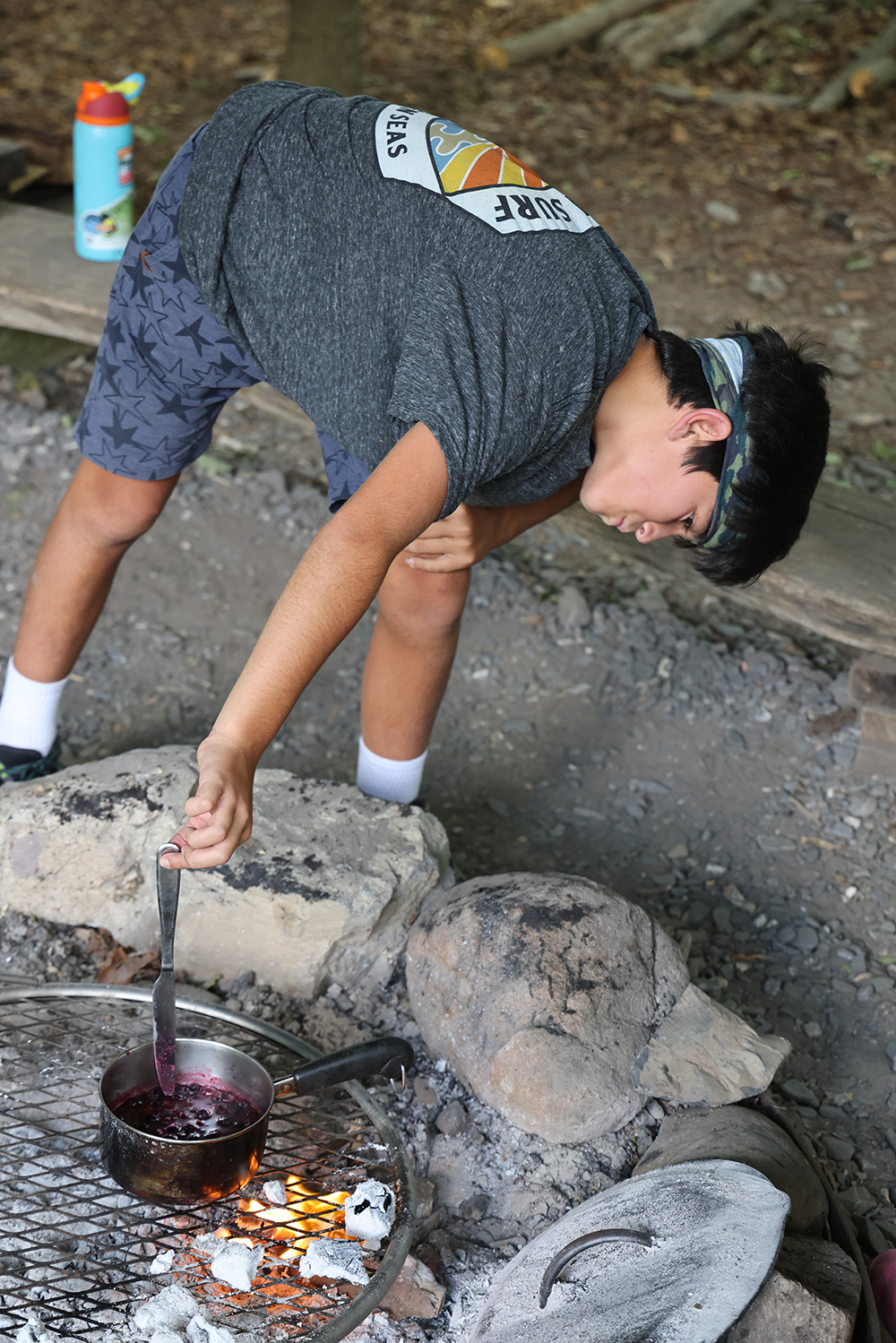 boy with dark hair leans over campfire to stir a small pot of blueberry sauce with a knife.