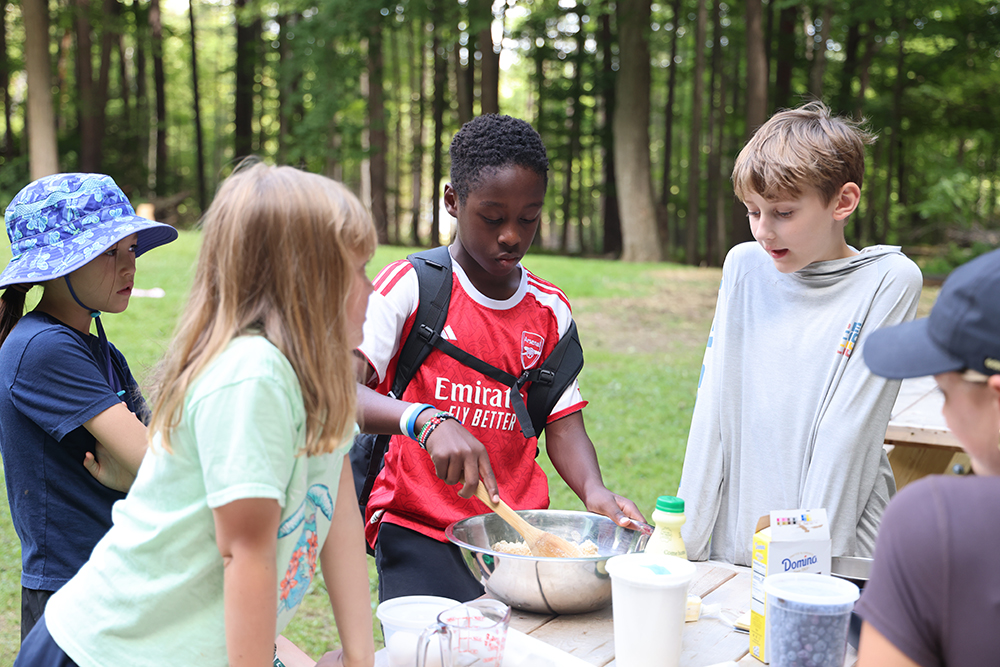 a small group of boys and girls standing at an outdoor picnic table while one student mixes batter in a bowl.