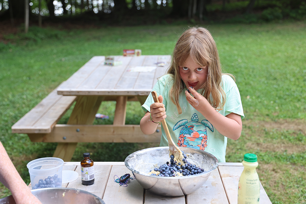 blond-headed girl stirring blueberry batter with a wooden spoon while sneaking some blueberries into her mouth and staring at the camera 