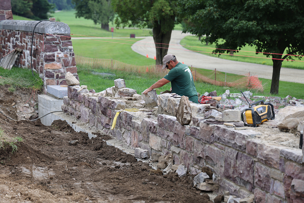 man in baseball cap lays a stone in a redstone wall being rebuilt, with green lawn, trees, and a winding dirt road behind him.