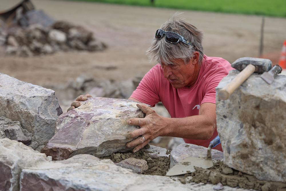 Man with short peppered hair grasps a big stone that he is setting in a stone wall.