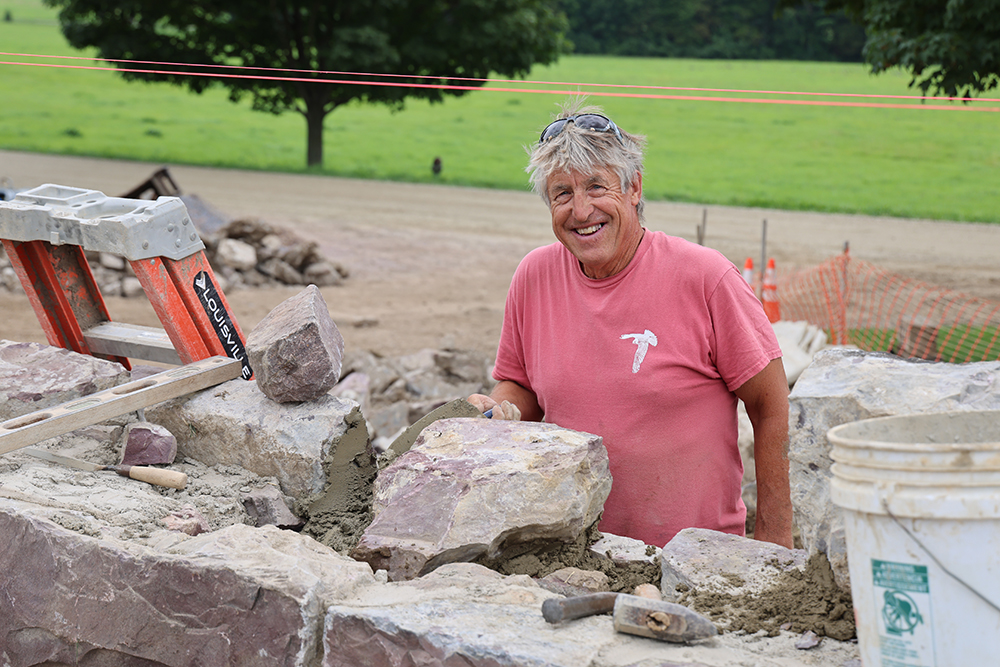 man with pepper hair looks at camera, holding a masonry trowel in his hand,stands behind stone wall that h