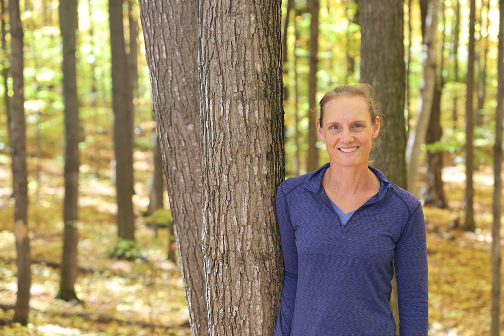 woman in blue shirt smiling at the camera, leaning against a tree with yellow woods in background
