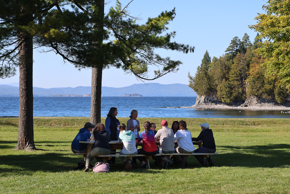 A dozen people talk around a picnic table with lake and mountains in distance