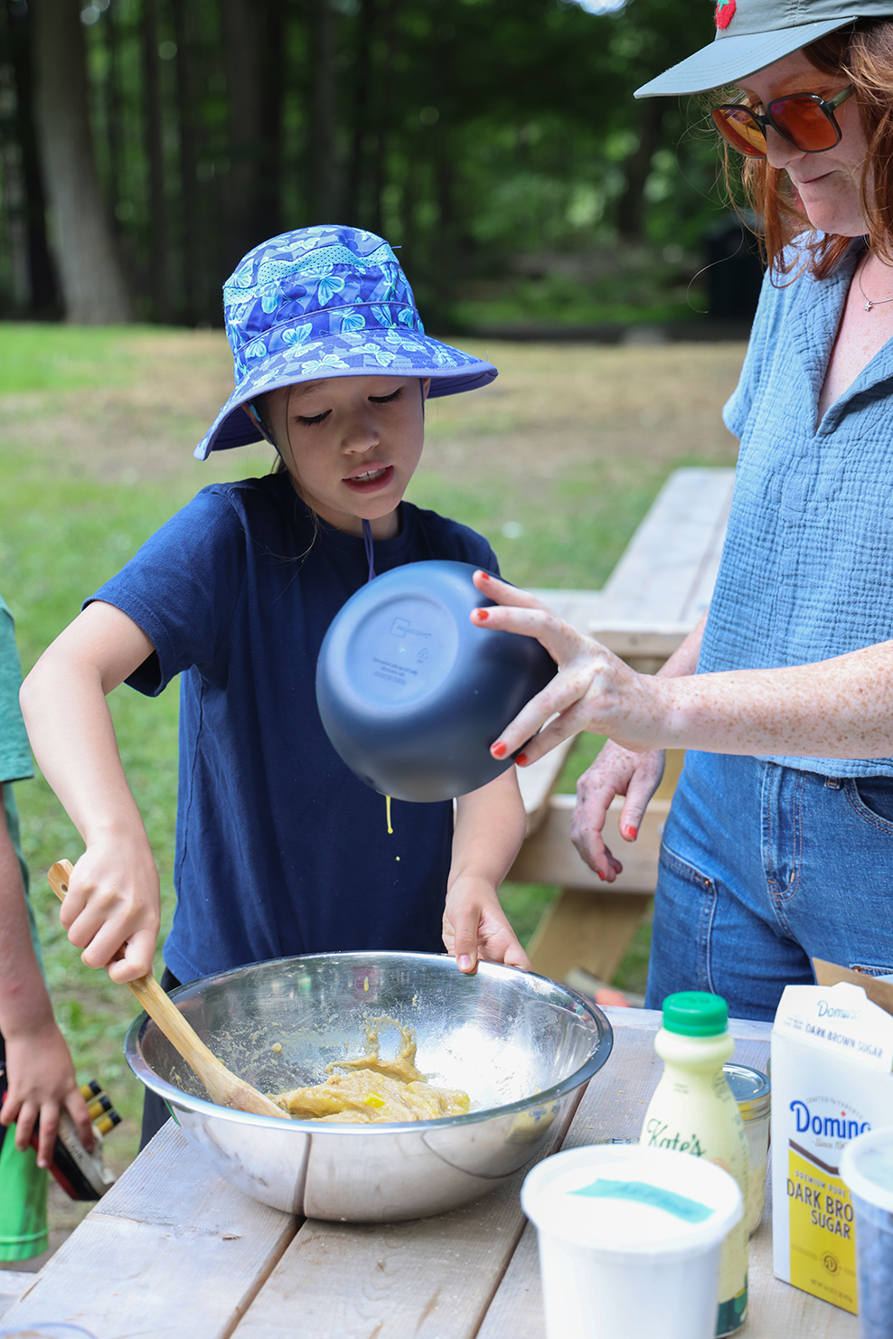 Woman in sunglasses pours egg from a small bowl into silver bowl on outdoor picnic table and girl in hat mixes egg into batter with wooden spoon..