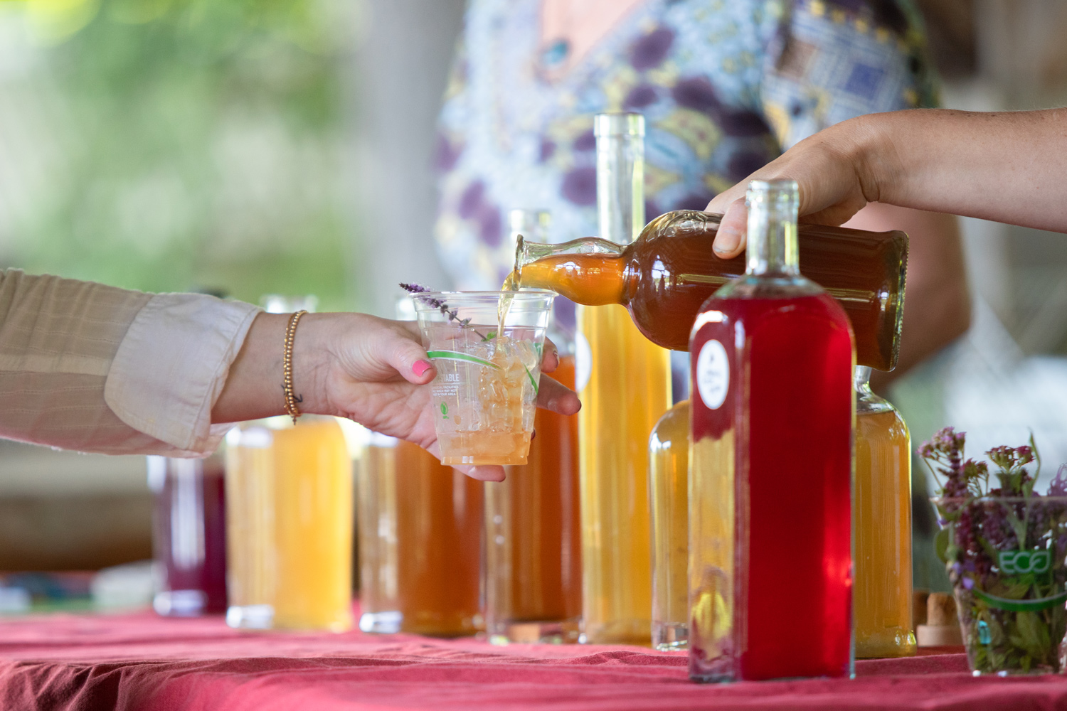 A person pours a beverage from a large bottle into a glass held by another person at a table with multiple bottles of colorful herbal syrups on a table.
