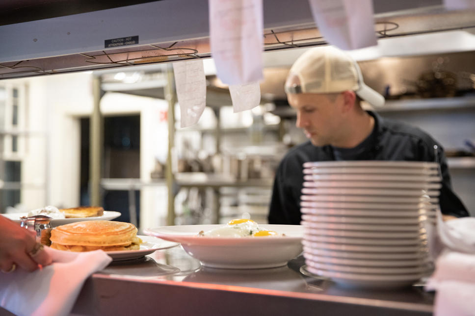 A man is seen through a kitchen pass cooking, plates of pancakes and eggs sit on the pass ready to be served