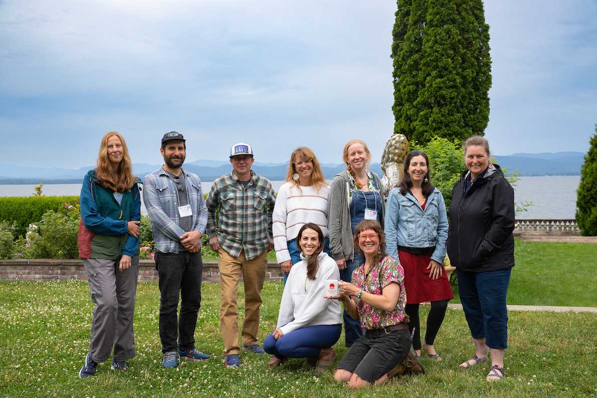 A group of people is gathered in a garden setting with a lake and mountains in the background