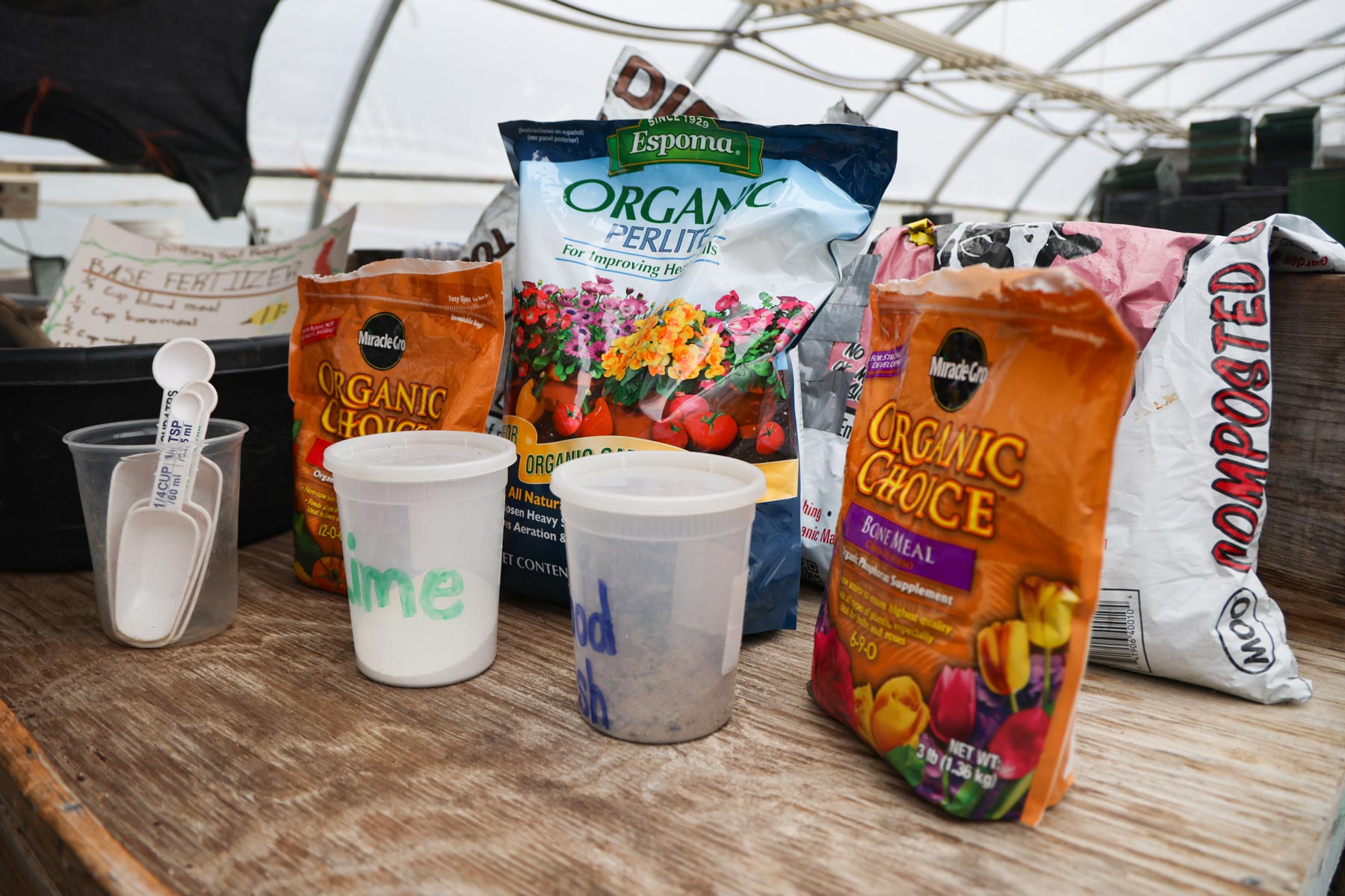 Bags of Espoma Organic Perlite and Miracle-Gro Organic Choice Potting Mix are on a wooden table in a greenhouse. Small containers labeled with different types of soil and scoopers are also visible.