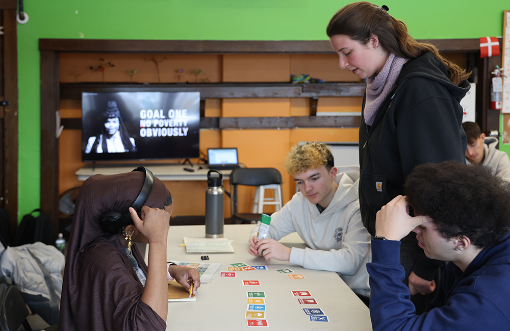 teens sit at a table looking at colorful cards representing the U.N. Sustainable Development Goals while woman teacher stands beside them