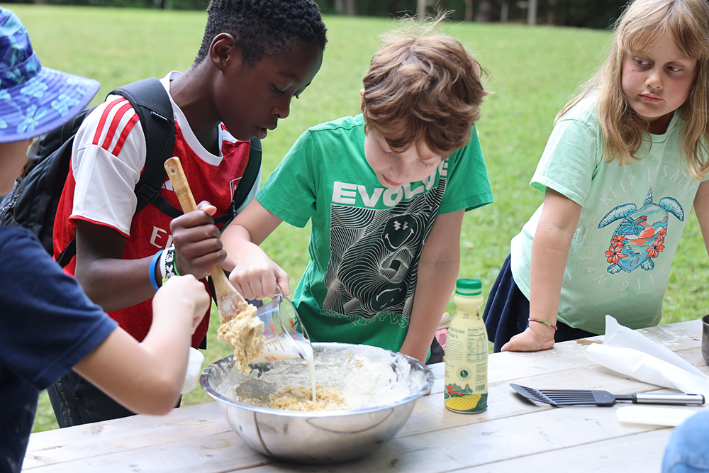 boy adds milk to silver bowl of batter while other boys mixes it in with wooden spoon and a girl looks on.