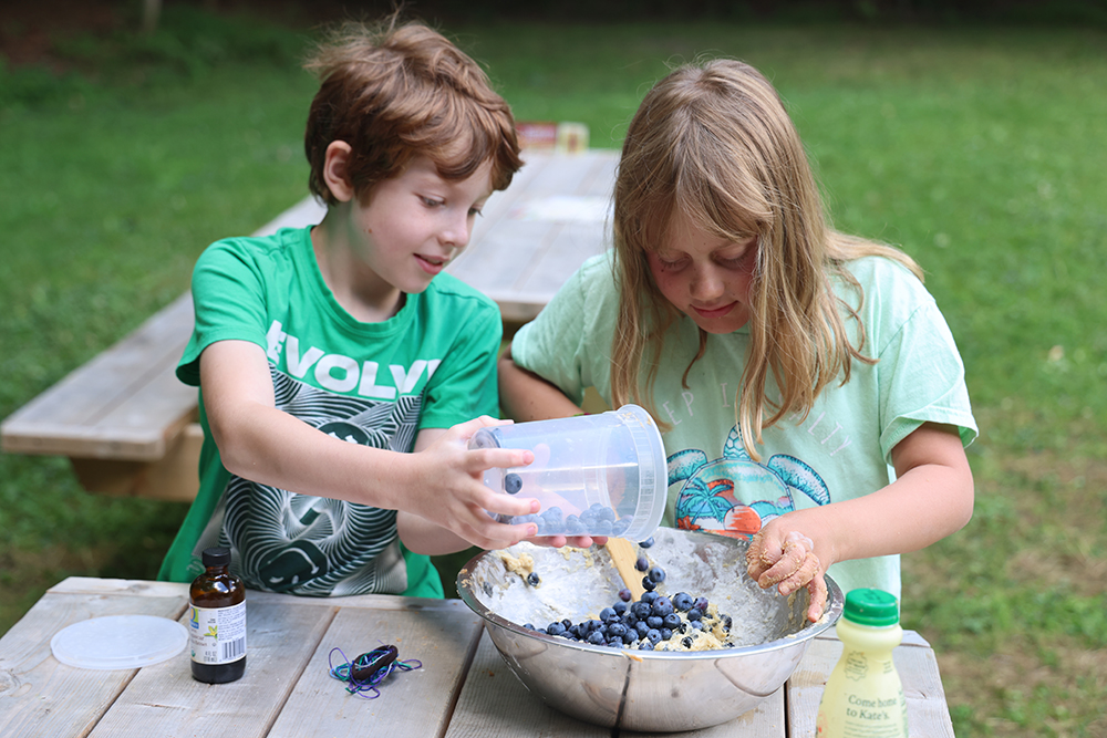 boy pours blueberries from plastic container into bowl of batter while girl mixes them in.