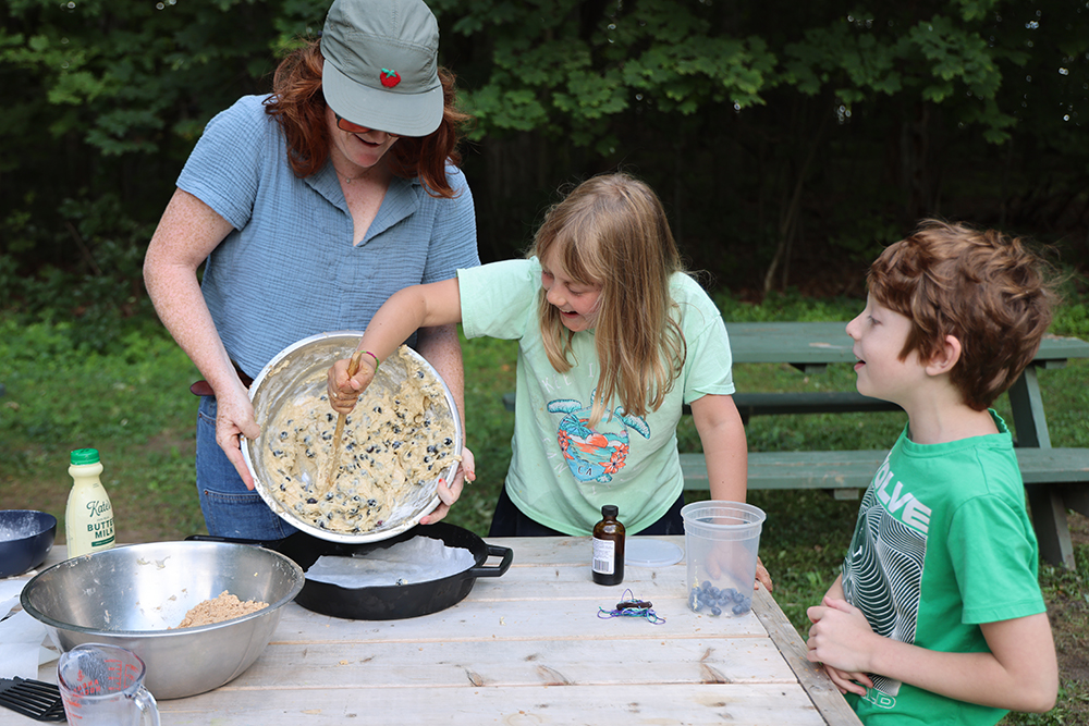woman holds blueberry batter over a skillet lined with parchment paper while girl scrapes batter into the skillet and boy looks on.