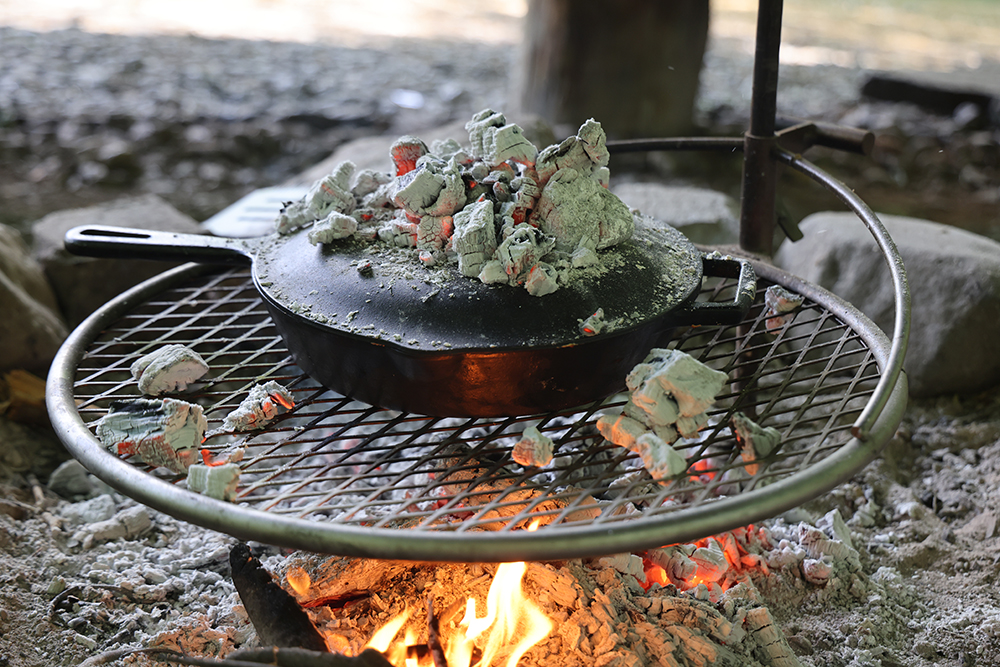 close up of a covered skillet on a rack over a campfire, with glowing coals sitting on top of the cover.