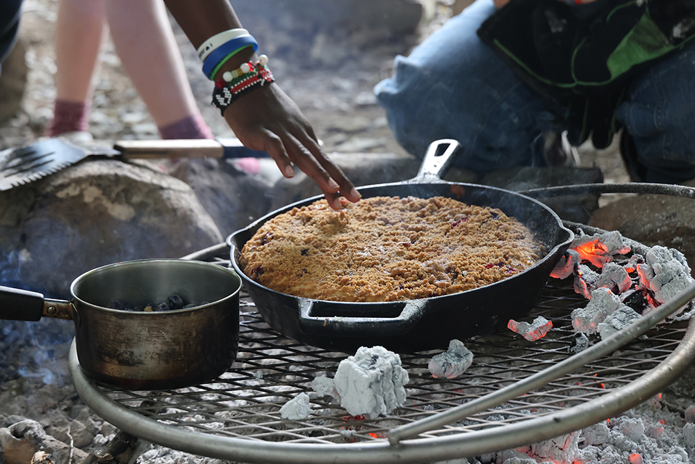 closeup of hand touching surface of coffee cake baking in skillet over a campfire.