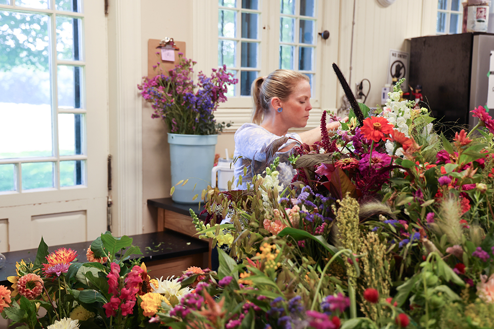 lots of cut flowers in foreground with woman in ponytail just behind the flowers arranging some