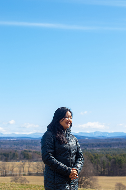 A portrait of Aimee Arandia Ostensen standing on the top of a hill in winter with sweeping views of rolling hills, mountains, and blue sky beyond