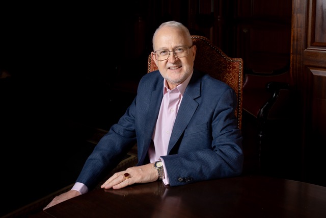 Photo of smiling man with white hair and glasses wearing a blue suit sitting at a dark wood table.
