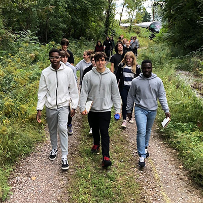 A group of high school students walk down a lush forested path with notebooks