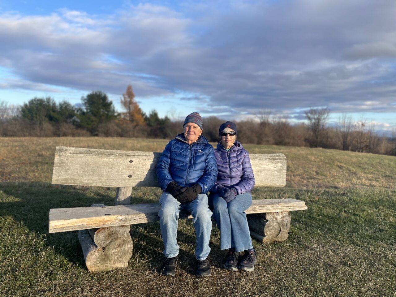 an older man and woman sit together on a rustic wooden bench surrounded by autumn grass. They are both in jeans, wearing winter coats, hats, and mittens.