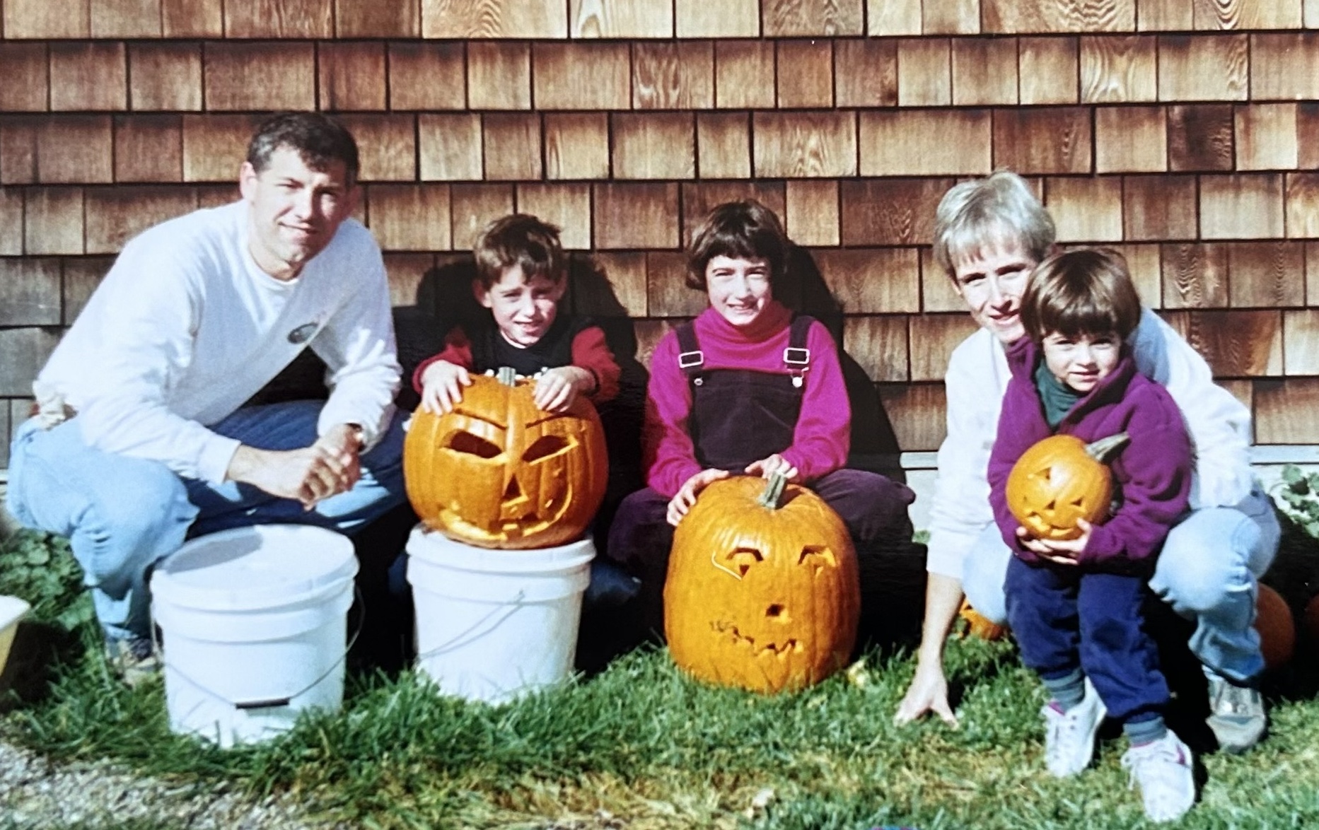 family of five squatting in front of shingled barn showing off three carved pumpkins. Left to right: Father, son, daughter, mom, youngest child