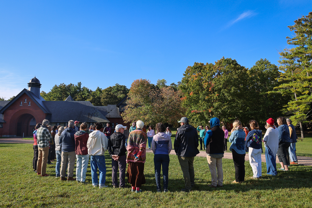 A large group of people stand in a circle outside of a brick building surrounded by trees