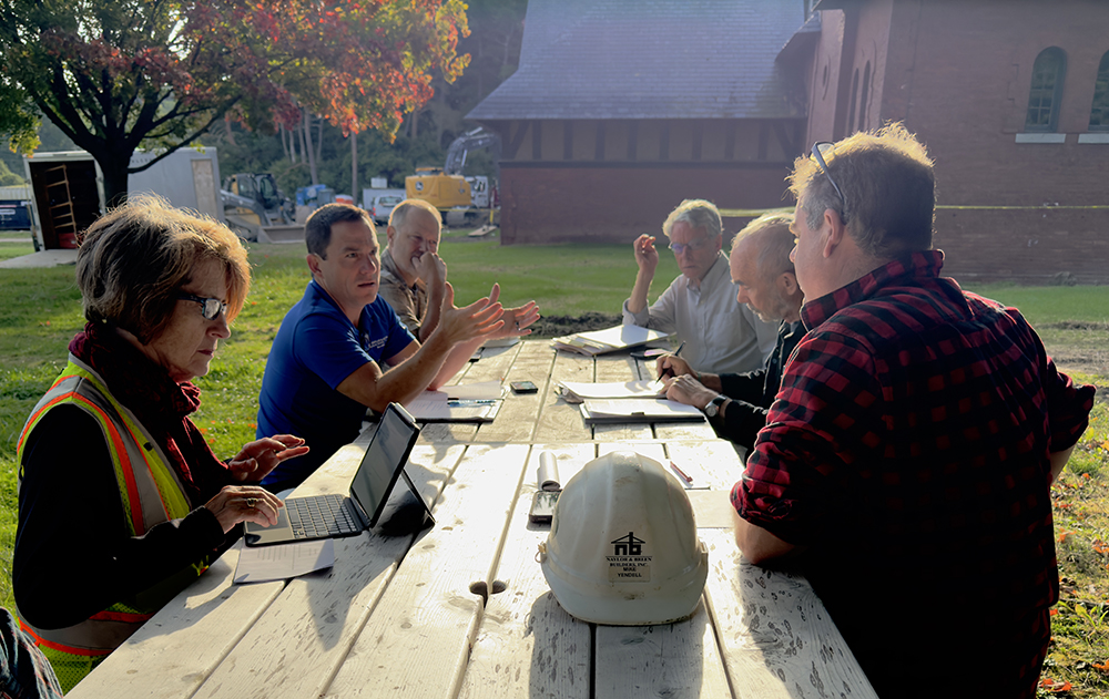 group of men and women gathered around a picnic table talking, some with computers and notebooks