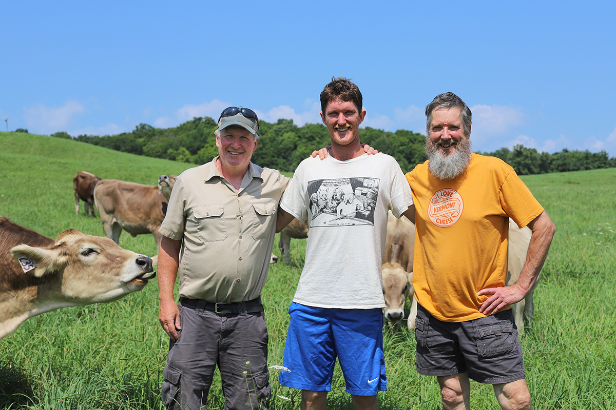 Sam, Gus, and Peter Dixon standing in a pasture together, with a cow trying to lick Sam's hand.