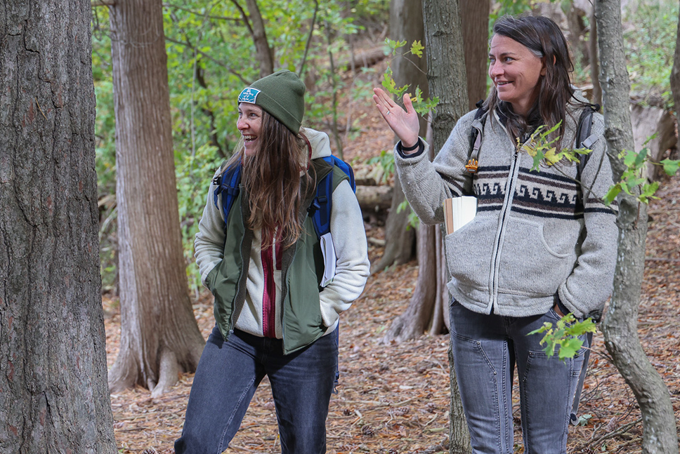 Two women smile while talking in a lakeshore forest