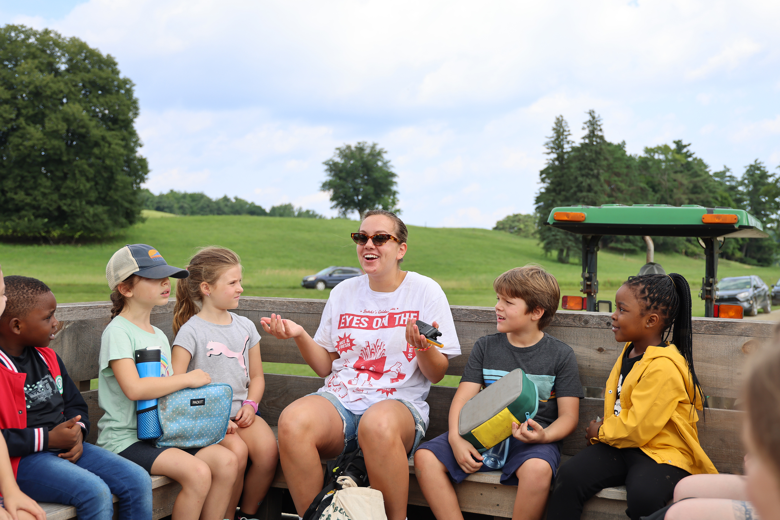 A woman rides in the a tractor pulled wagon with a dozen young summer campers on a farm