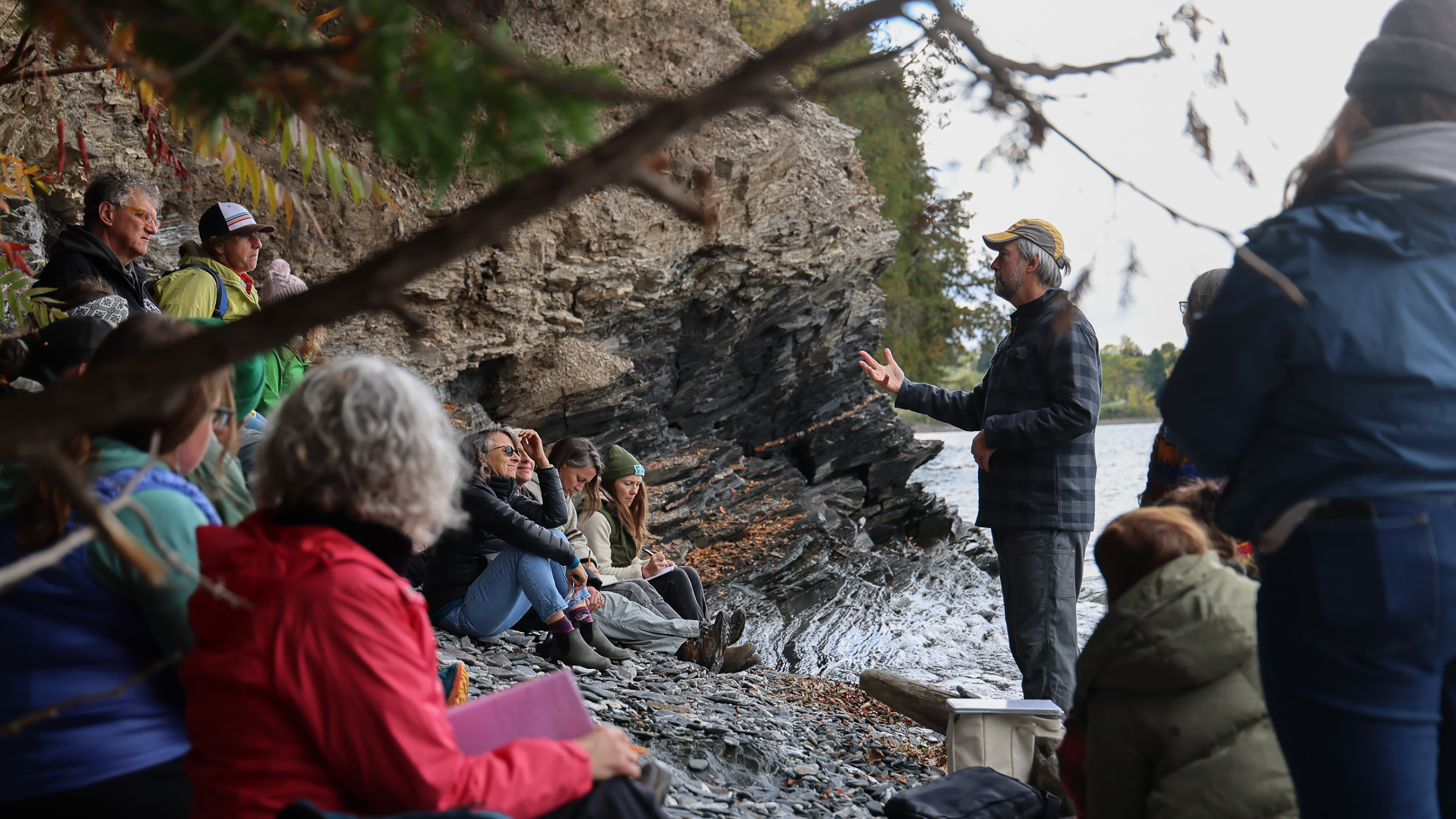 A dozen adults sit and stand along a rocky lake shoreline in fall, listening to an instructor