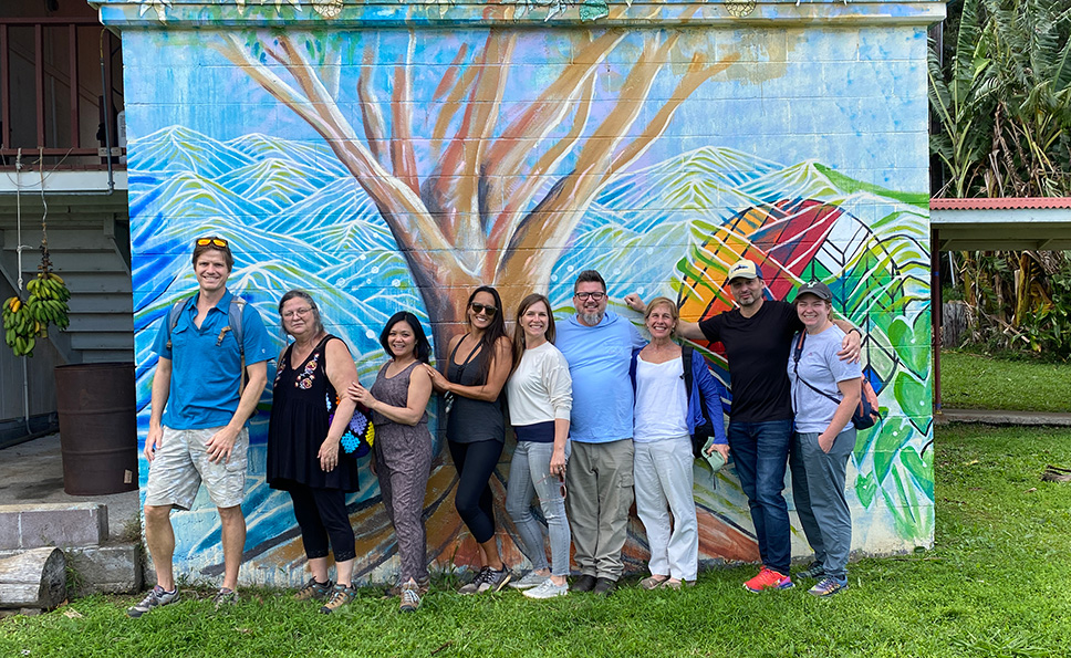 Ten adults pose and smile in front of a large colorful botanical mural on a brick wall. They stand on lush green grass