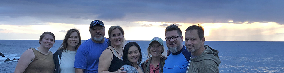 Eight people stand on an ocean overlook in Hawai'i, with a cloudy sunset and dark ocean behind them, smiling