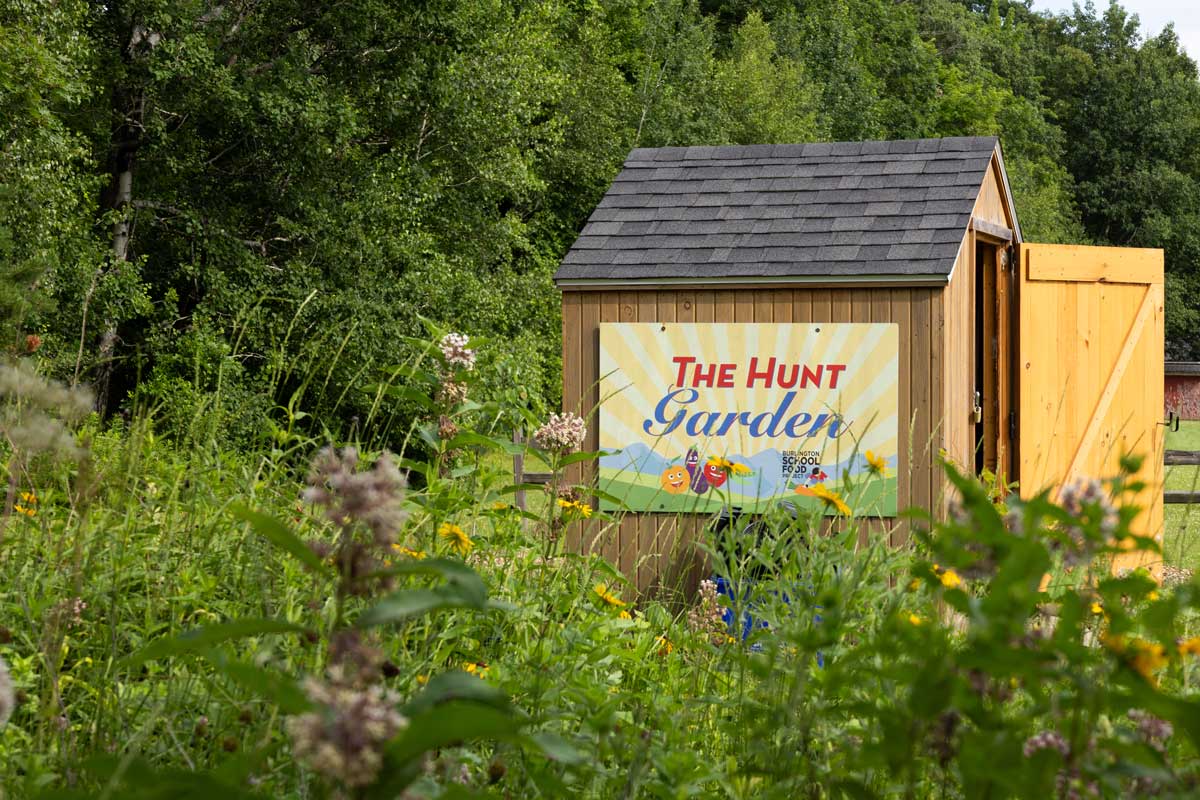 Small wooden shed featuring a sign that reads "The Hunt Garden," surrounded by lush greenery and wildflowers. 