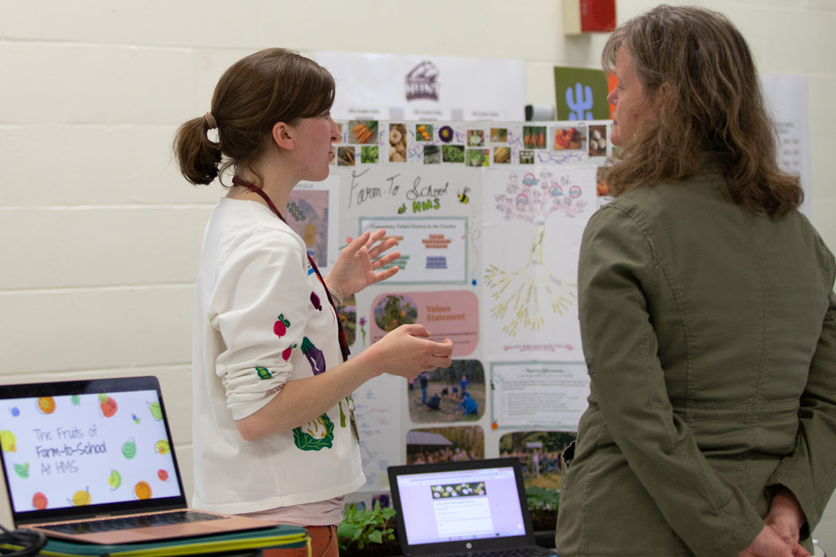 Two individuals are engaged in conversation in front of a display board titled "Farm to School" with various images and text. Nearby, a laptop shows "The Fruits of Farm-to-School at MHS" on the screen.
