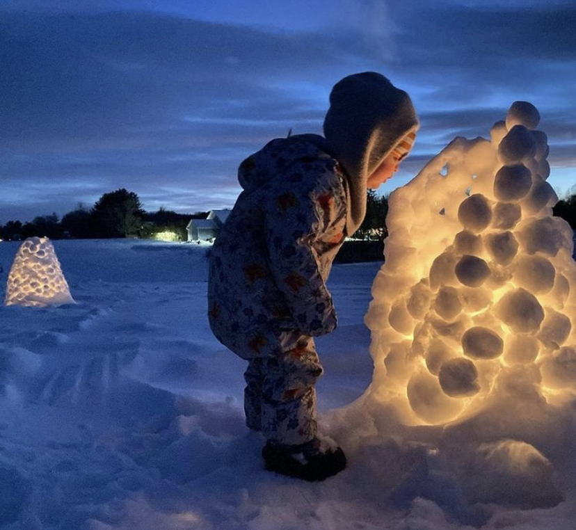 Young girl in snowsuit peers into a lit snow candle at twilight