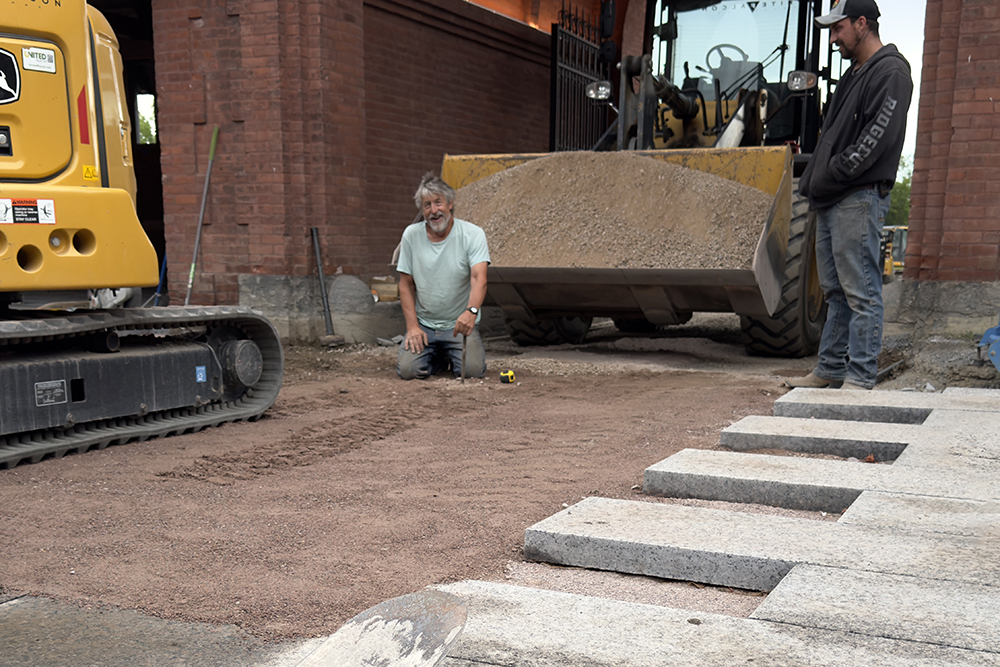 man kneels in packed gravel, surrounded by heavy machinery to pack and add more gravel. Granite pavers are in the foreground.