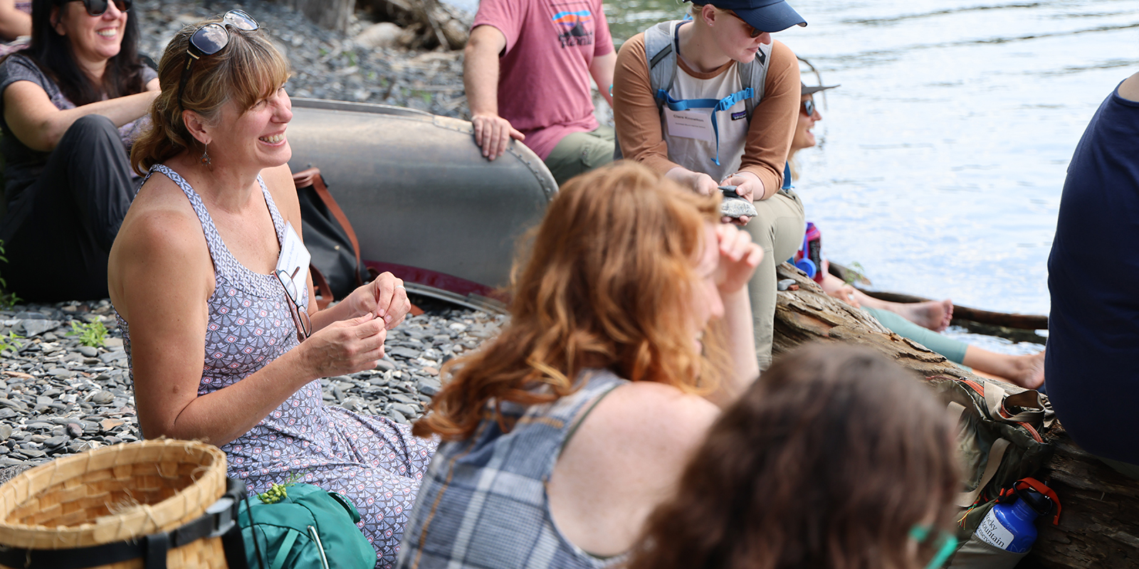 A group of adults smile while sitting on a rocky lakeshore listening to a person speak