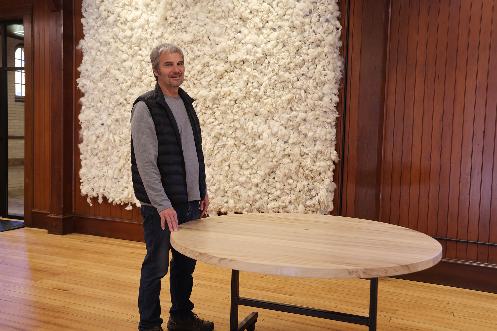 man with salt-and-pepper short hair stands in jeans next to a table with a round wooden top. A wall decoration of sheep's wool hangs behind him