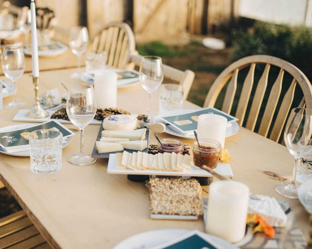 Elegant outdoor dining table setup featuring an array of plates, glasses, and candles, with assorted cheeses, fruits, and condiments arranged for a meal.