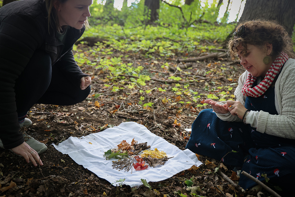 Two teachers sit on the forest floor making art from natural materials