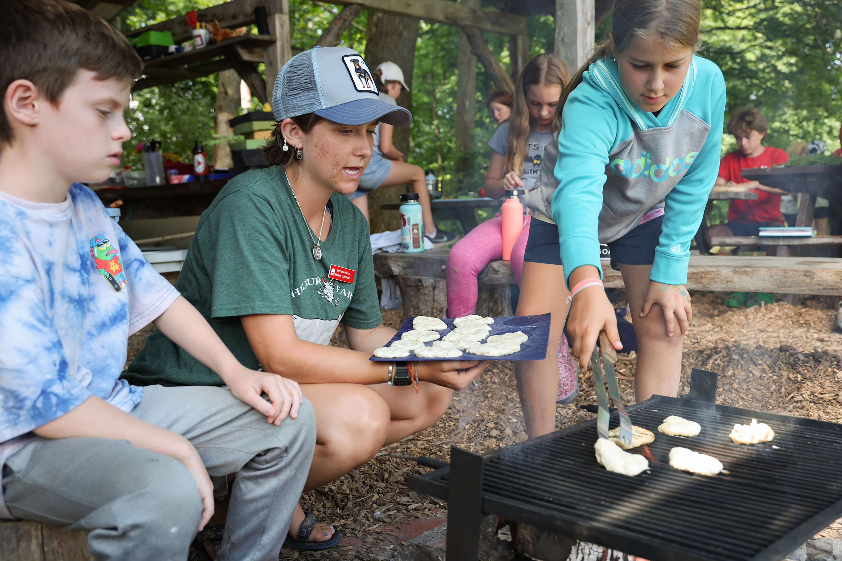 A woman flips flatbread over an open campfire with children on either side under an outdoor pavilion