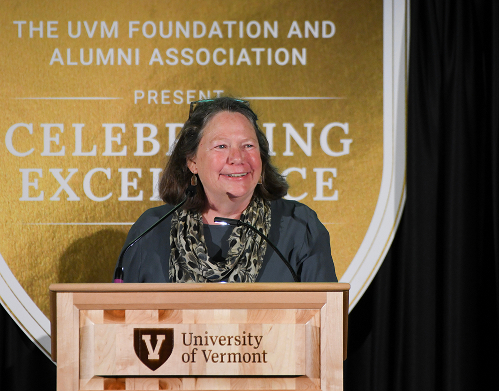 Megan Camp, a woman with shoulder length hair, stands at a UVM podium making a speech
