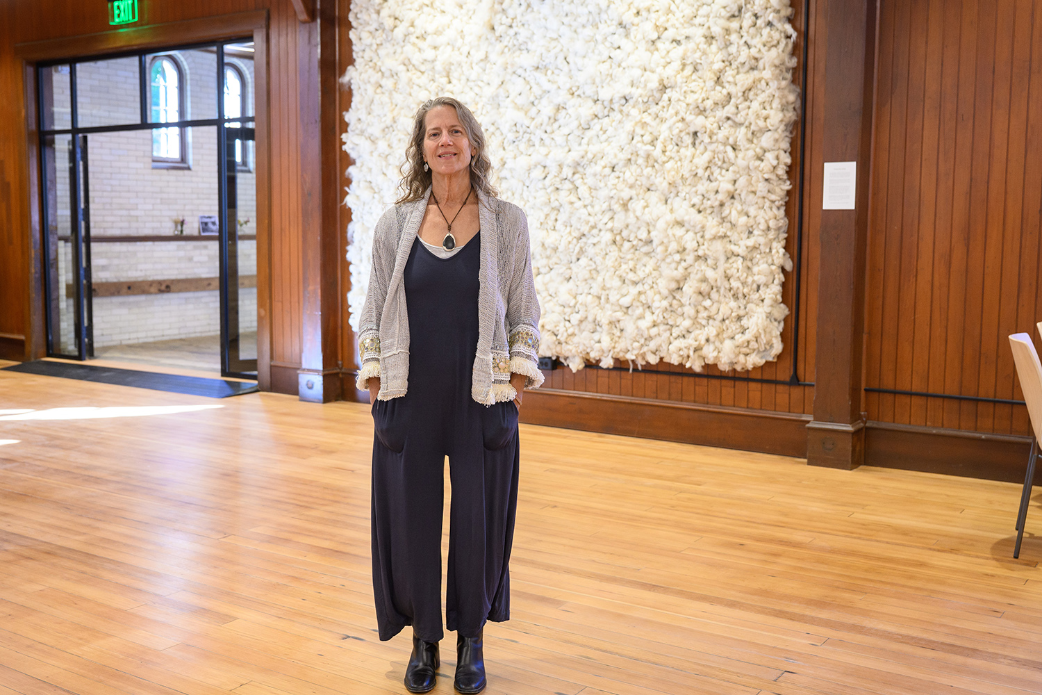 woman in gray sweater over long black pantsuit smiles at camera as she stands in front of wall art made of tufts of white sheep's wool