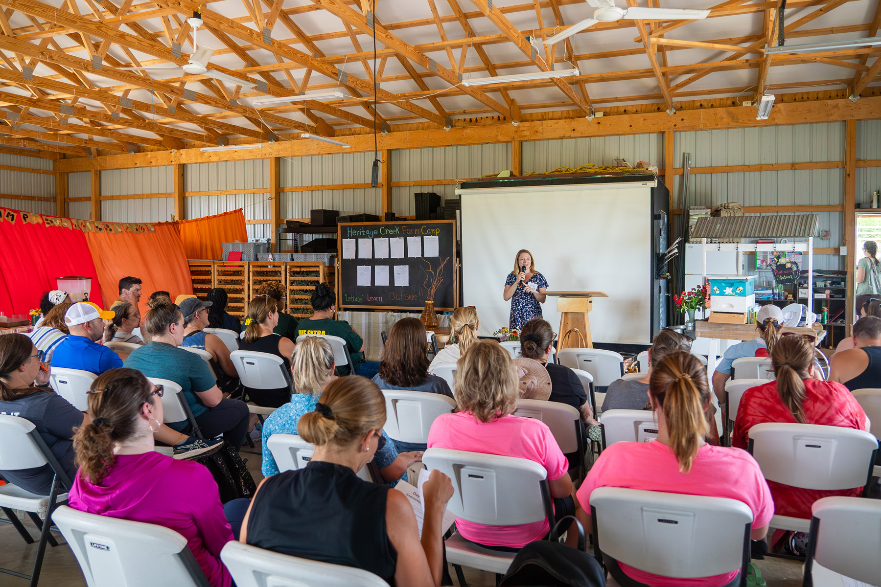 A person giving a presentation at a podium to an audience seated in a hall with educational materials displayed.