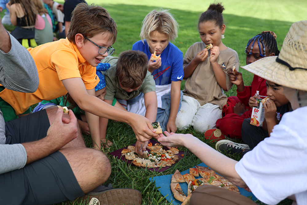 A group of young children enjoy a pizza together on a grassy lawn